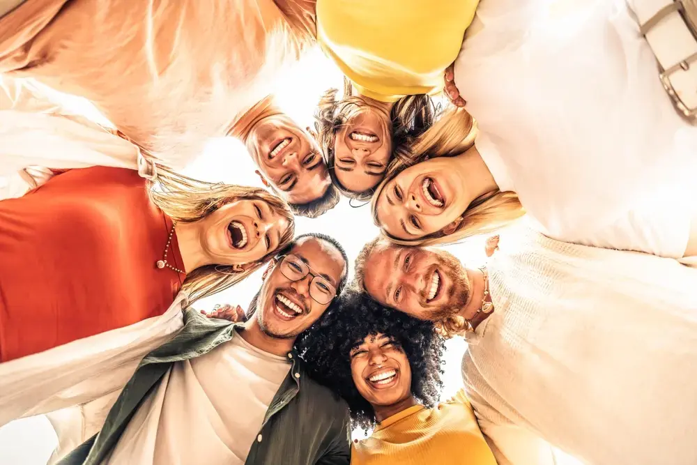 A diverse group of seven friends stands in a circle, smiling and laughing, viewed from below against a bright sky. Their joyful connection highlights the sense of togetherness and employee wellbeing.